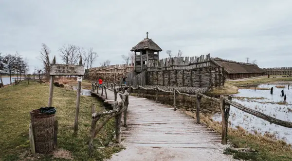 Drewniany mostek prowadzi do grodu z wieżą obronną i chatami krytymi strzechą.