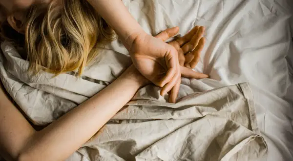 Blonde Haare auf einem Kissen, Arme schlafen nachts ein, Hände ruhen auf der Brust, umgeben von zerknitterter Bettwäsche.