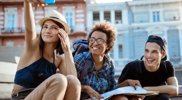 Tres amigos sonríen mientras se toman una selfie en la calle, planeando cómo conocer gente para viajar y explorar la ciudad.