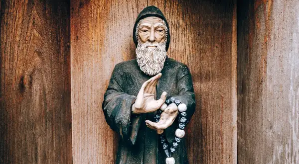 Posąg mnicha z brodą i szarbel uzdrowienia w dłoniach, na tle drewnianej ściany.