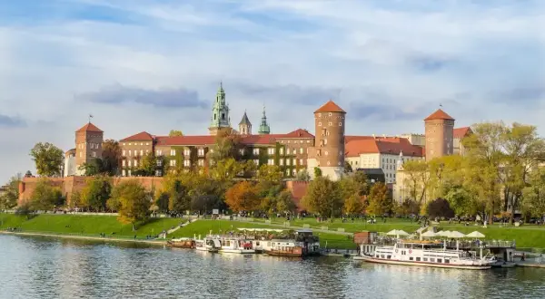 Malownicze miasto nad Wisłą z Wawelem w jesiennej odsłonie. Statki wycieczkowe cumują przy brzegu, a na trawniku odpoczywają spacerowicze.