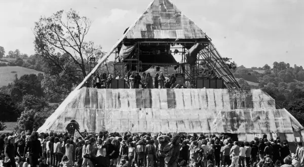 A massive pyramid stage hosts performers at the first Glastonbury Festival. A huge crowd gathers below, marking a historic moment in music history.