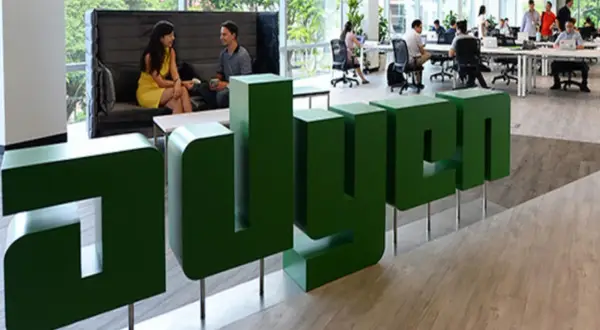 Zielone, przestrzenne litery z napisem "adyen" w nowoczesnym biurze. W tle ludzie pracują przy biurkach i rozmawiają.