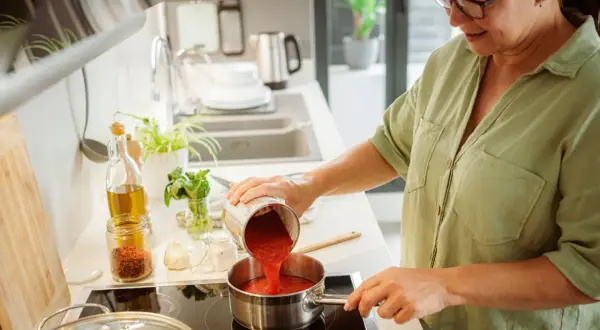 Mujer vertiendo tomate triturado en una olla para espesar una salsa. Ingredientes como aceite, ajo y especias a la vista.