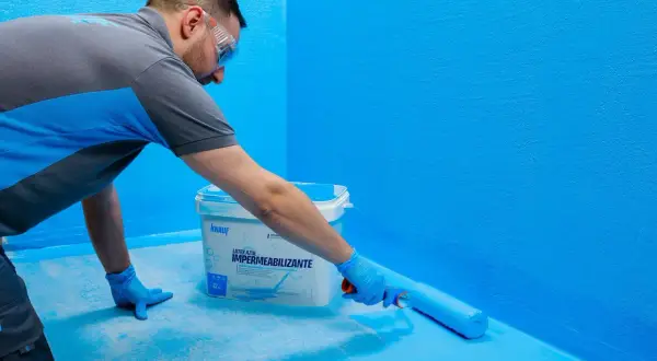 Hombre aplica látex azul con rodillo, creando sistemas de impermeabilización en paredes y suelo.