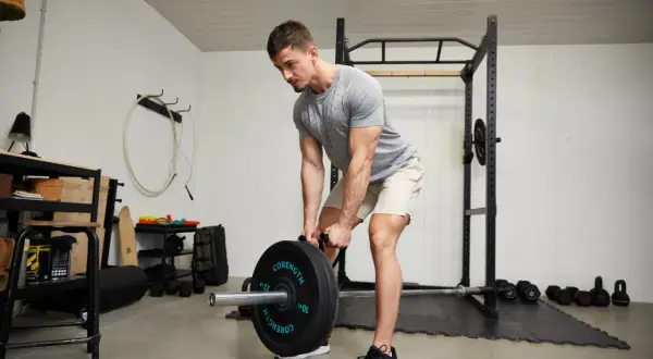 Homme musclé en buste penché, soulevant une barre chargée de poids dans une salle de sport à domicile.