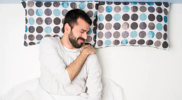 Uomo a letto con dolore alla spalla, dormire con le braccia alzate non è comodo.