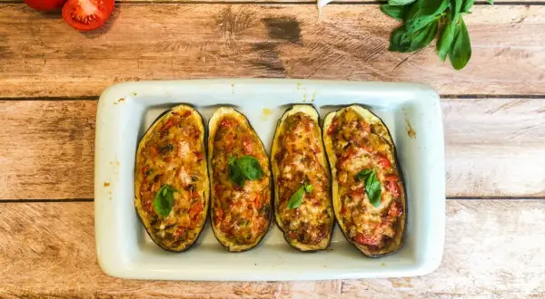 Quatre moitiés d'aubergines farcies, gratinées et garnies de basilic, dans un plat blanc sur fond de bois.