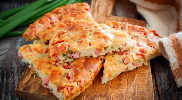 Pyszna tarta z kiełbasą, pokrojona na kawałki, leży na drewnianej desce. Obok zielona cebulka i ręcznik.