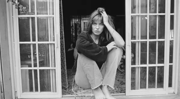 Jane Birkin mit typischen 70er Frisuren, lässig auf einem Stuhl sitzend, vor einer Tür mit Sprossenfenstern.