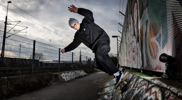 Martin Ander gör ett trick på skateboard i en betongpark med graffitiväggar och järnvägsspår i bakgrunden.