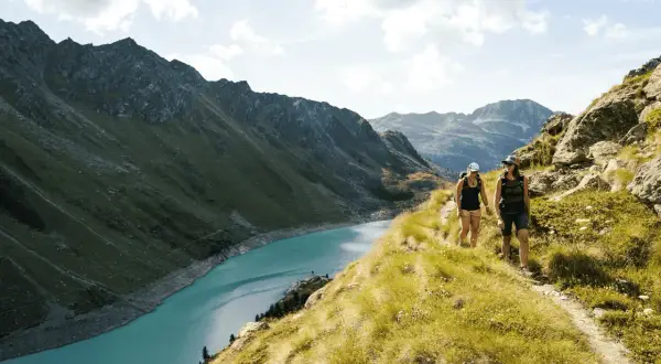 Två personer vandrar på en bergstig med en turkos sjö och dramatiska berg i bakgrunden. En perfekt dag för att vandra i Schweiz.