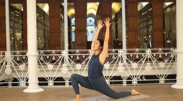 Mujer en postura de yoga, con brazos extendidos hacia arriba, practicando dharma yoga en una sala con estanterías de libros.
