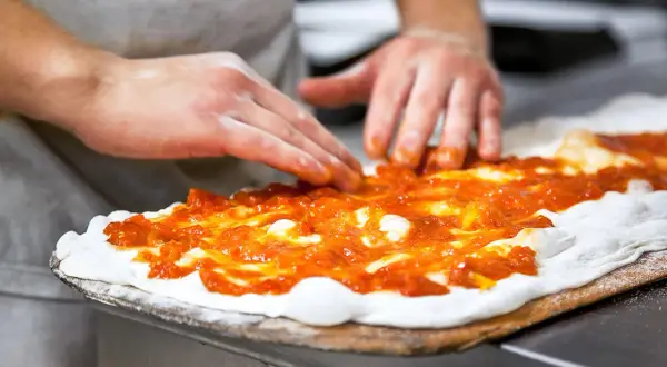 Händer sprider tomatsås på pizzadeg. En del av en matlagningskurs i att baka pizza.
