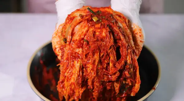 Ręce w białych rękawiczkach trzymają głowę kapusty pekińskiej pokrytą czerwoną pastą. To właśnie co to jest kimchi – tradycyjna koreańska kiszonka.