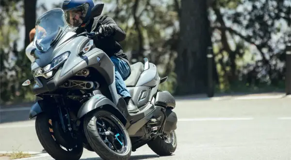 Hombre en casco azul conduce un triciclo Yamaha Tricity 500 por una carretera arbolada, inclinándose en una curva.