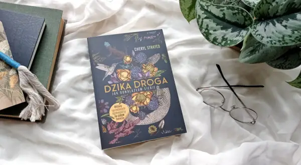 Okładka książki "Dzika droga" z motywami roślinnymi i zwierzęcymi, obok okulary i inne książki.