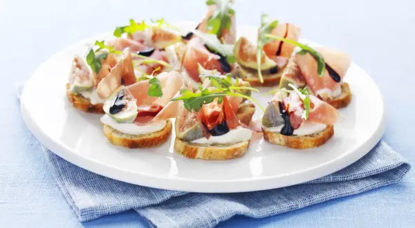 Köstliche italienische Crostini mit Prosciutto, Feigen, Mozzarella und Rucola auf einem weißen Teller.