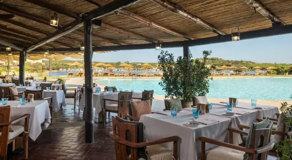 Tavoli apparecchiati sotto un tetto di paglia, con vista sulla piscina e sulla spiaggia del **hotel Cala di Volpe**.