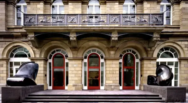 Vor dem Museum Wuppertal stehen zwei abstrakte Skulpturen. Die Fassade des Gebäudes ist aus hellem Stein, mit roten Türen und verzierten Balkonen.