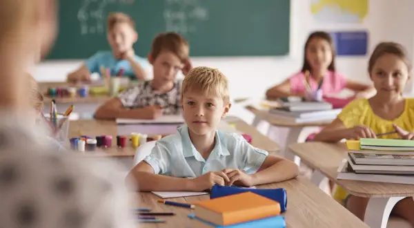 Dzieci siedzą w klasie przy ławkach, przed nimi książki i przybory.