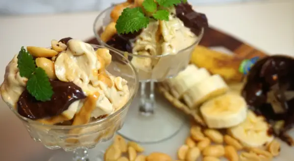 Två glas med nyttig glass, toppad med choklad, jordnötter och mynta. Bananskivor och jordnötter ligger bredvid.