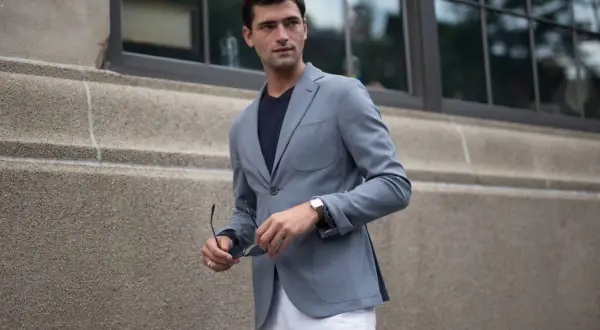 Ein Mann mit einem grauen Sakko kombiniert es lässig mit einem dunklen T-Shirt und weißen Hosen. Er hält eine Sonnenbrille in der Hand.