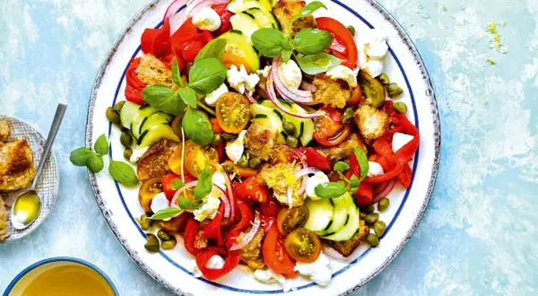 Ensalada de burrata vibrante con tomates, pepino, pimientos rojos, pan tostado y alcaparras, adornada con albahaca fresca.