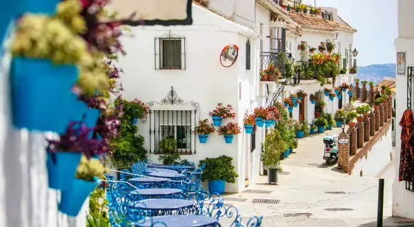 Callejuela encantadora en uno de los pueblos costeros de Málaga, con fachadas blancas, macetas azules y mesas de terraza.