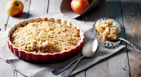 Un délicieux crumble aux pommes tout juste sorti du four, servi dans un plat rouge, avec une portion sur une cuillère ornée.