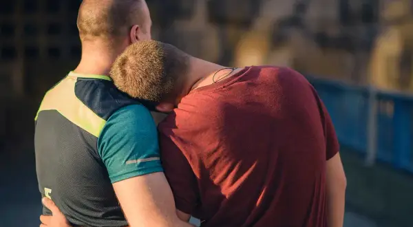 Två män omfamnar varandra. Den ena vilar huvudet mot den andras axel, en bild av en varaktig relation.
