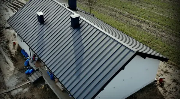 Nowy, ciemnoszary dach z blachy na domu jednorodzinnym. Widać kominy i otoczenie budowy. Dobry koszt dachu to podstawa.