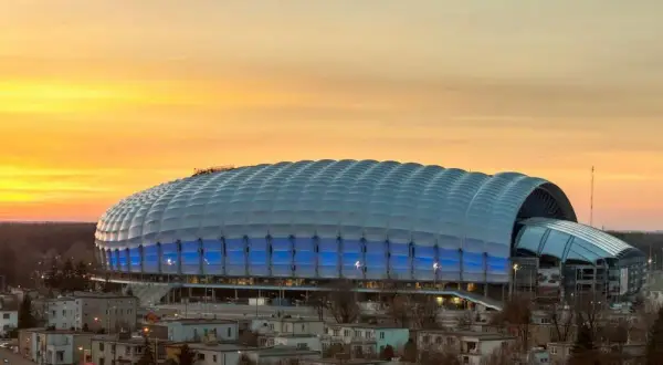 Stadion miejski w Poznaniu o zachodzie słońca, z niebieskimi światłami i charakterystycznym, falującym dachem.