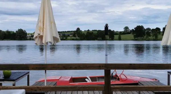 Gemütliches Restaurant am See mit rotem Tretboot und Sonnenschirmen. Die Uferlandschaft ist grün und hügelig.