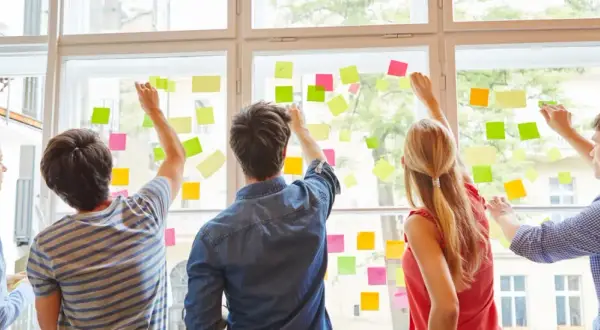 Fünf Personen planen an einem Fenster mit bunten Haftnotizen. Sie arbeiten an ihrer kaufmännischen Ausbildung nebenberuflich.