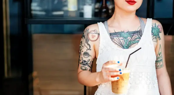 Junge Frau mit vielen Tattoos, darunter ein Vogel und ein Auge. Sie hält einen Eiskaffee. Ein Tattoo ab 16, das ihre Persönlichkeit unterstreicht.