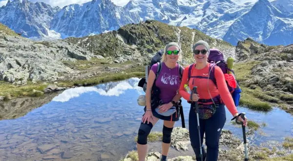 Dwie kobiety cieszą się trekkingiem wokół Mont Blanc. Mają plecaki i kije trekkingowe, a w tle widać ośnieżone szczyty.
