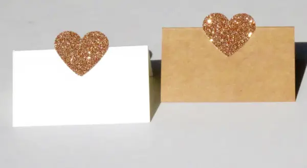 Segnaposto nozze d'oro fai da te: due cartoncini, uno bianco e uno kraft, decorati con cuori glitterati color oro.