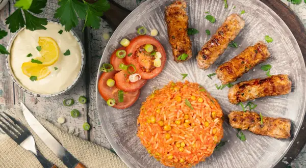 Saftige cevapcici im backofen, serviert mit Reis, Tomaten und einer cremigen Sauce.