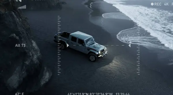 Un camion du futur gris, vu du ciel, roule sur une plage de sable noir. Les vagues de l'océan s'écrasent sur le rivage.