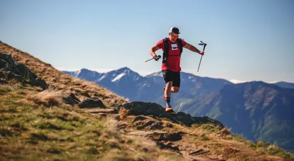 Ein Mann im roten Shirt und schwarzer Hose läuft mit Stöcken einen Bergpfad entlang. Sein intensives anaerobes Training fordert ihn in der atemberaubenden Berglandschaft.