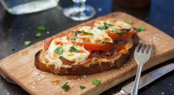 En härlig tallrik med varma mackor, toppad med smält ost, tomater och färska örter. Serveras på en träskärbräda med bestick.