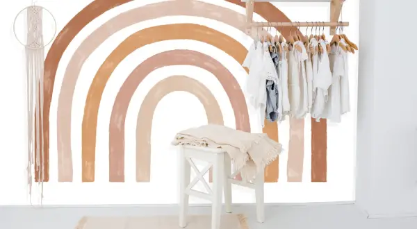 Un arc-en-ciel aux tons terreux est peint sur un mur blanc, orné d'une penderie d'enfants et d'une décoration murale en macramé.