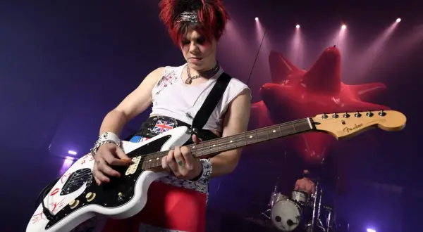 Yungblud gra na gitarze podczas koncertu w Warszawie. W tle czerwona, dmuchana instalacja i perkusista.