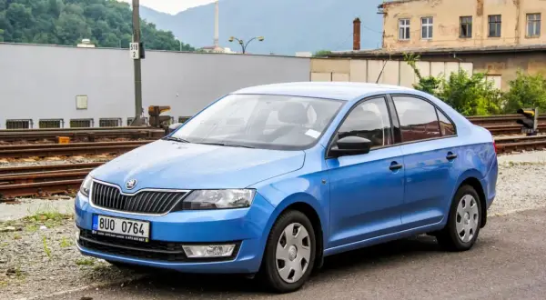 Niebieski sedan Skoda Rapid stoi przy torach kolejowych. W tle budynki i góry.