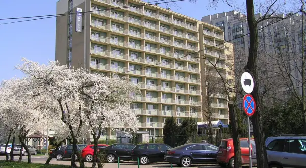 Hotel Felix Warszawa w otoczeniu kwitnących drzew. Widać parking z samochodami i budynek hotelu z balkonami.