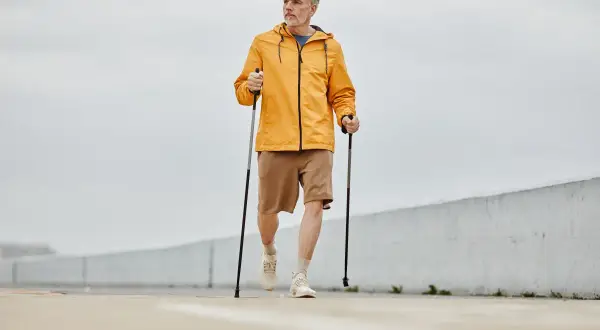 Mężczyzna w żółtej kurtce i brązowych spodenkach wykonuje rozgrzewkę przed nordic walking, trzymając kije.