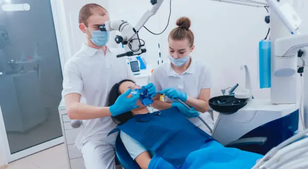 Dentysta pod mikroskopem wykonuje ekstyrpację miazgi u pacjentki, asystuje mu pomoc dentystyczna.
