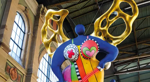 Silhouette d'une Nana colorée de Niki de Saint Phalle, avec des ailes dorées, suspendue dans un grand hall.