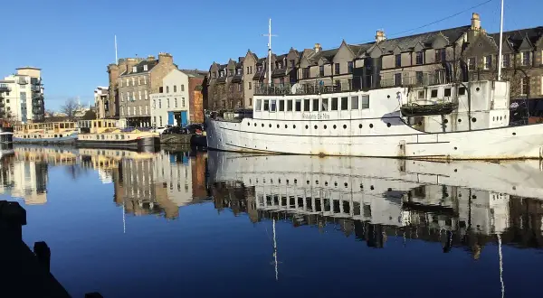 Barcos y edificios históricos reflejados en el agua en Leith, Edimburgo. El "Ristorante De Niro" en un barco blanco destaca.